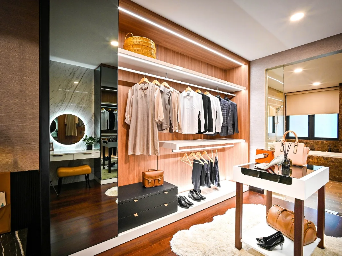 Modern walk-in closet, crafted by custom closet designers, featuring organized clothing on hangers, shelves with bags, a dressing table with mirror, and a bench on a wooden floor with a white rug.