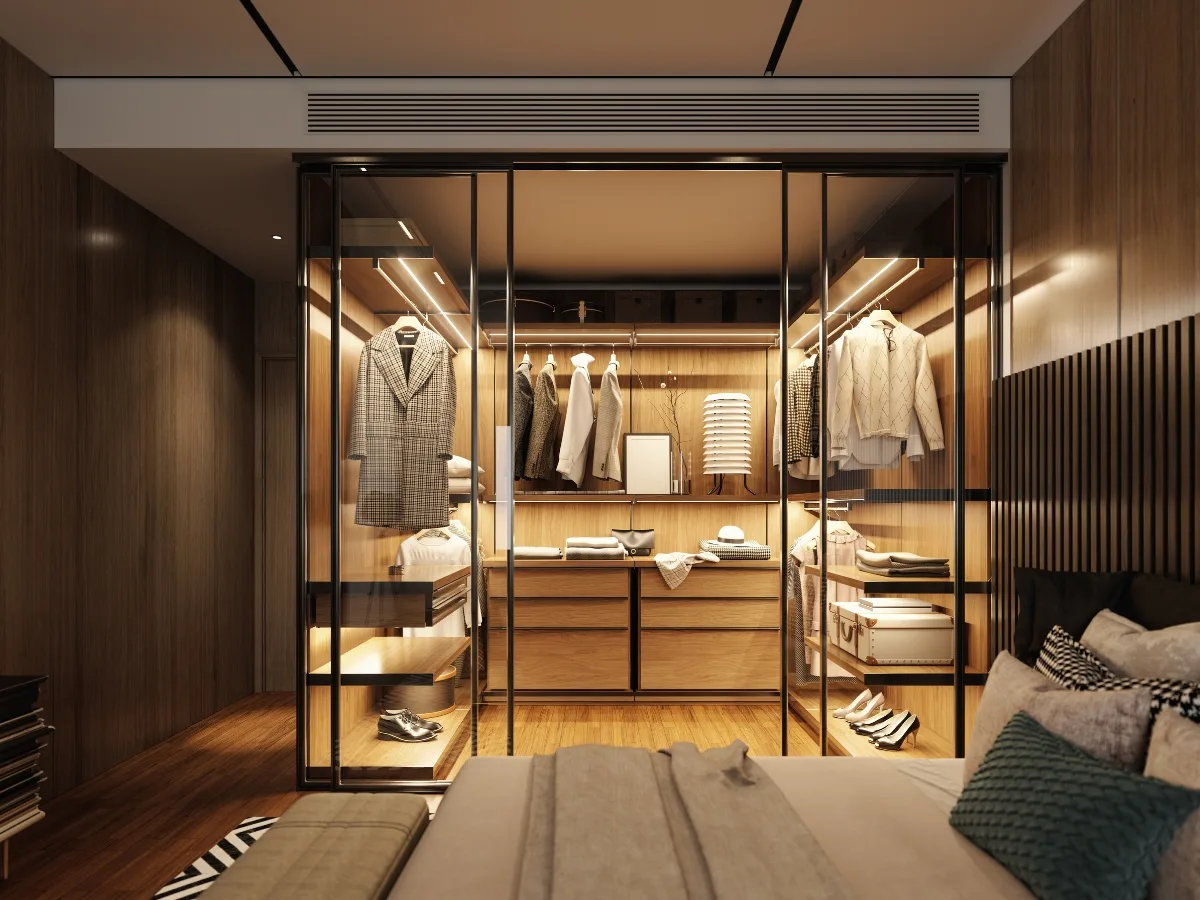 Modern walk-in closet with glass doors, wooden shelves, hanging clothes, and drawers&mdash;expertly crafted by custom closet designers&mdash;adjacent to a neatly made bed in a stylish bedroom.