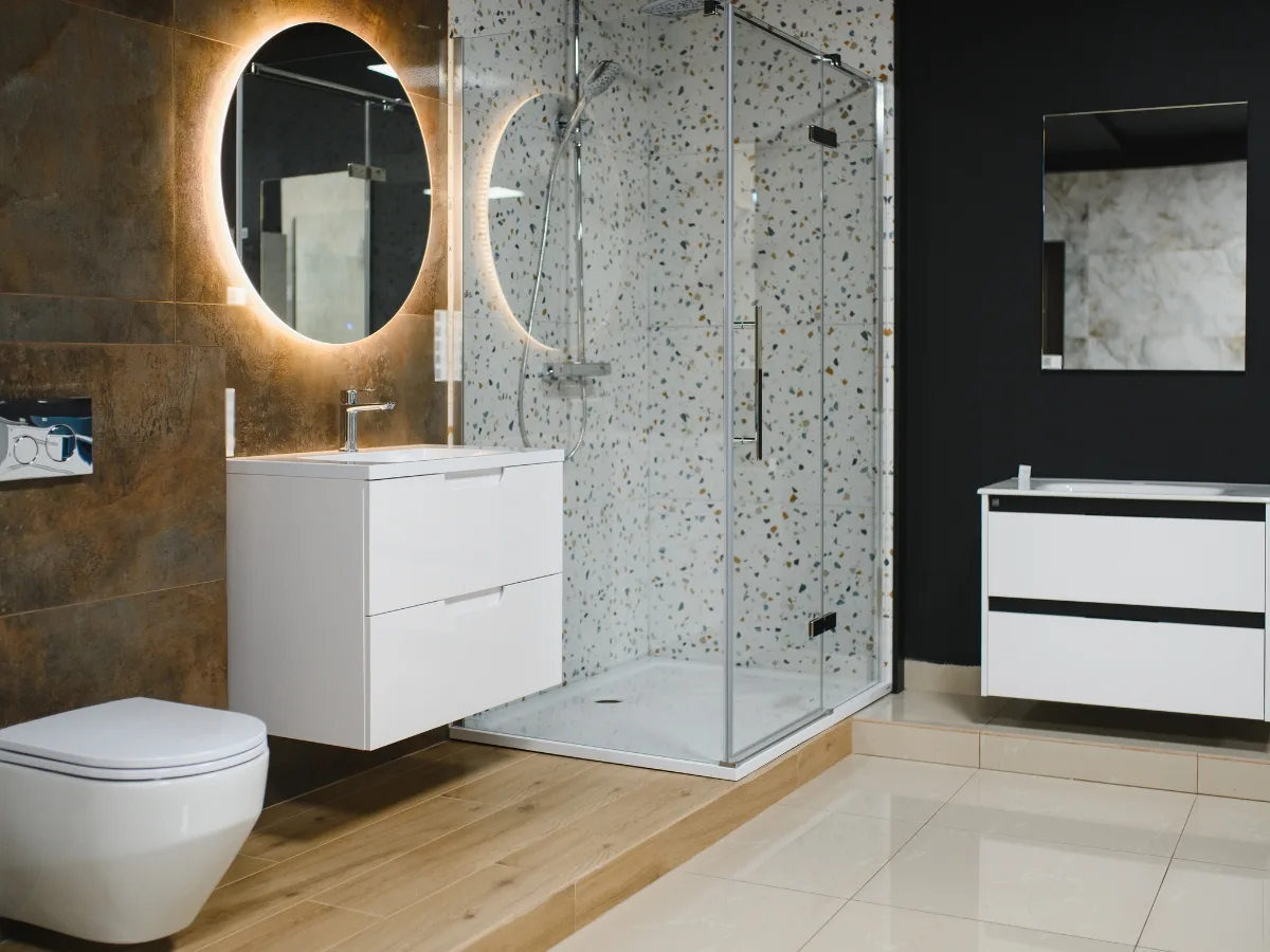 Modern bathroom with a wall-mounted toilet, floating vanities, round backlit mirror, glass shower enclosure, wood and tile flooring, and patterned shower wall tiles—perfect for Buckhead GA luxury bathroom remodeling.