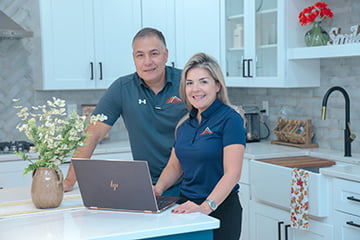 Two people stand in a modern kitchen, both smiling at the camera. One is using a laptop on the counter. White cabinets and a vase of flowers are visible in the background.
