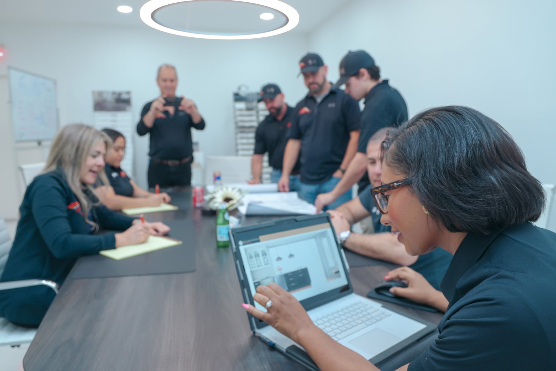 A group of people in black shirts sit and stand around a conference table during a meeting; one person presents information on a laptop.