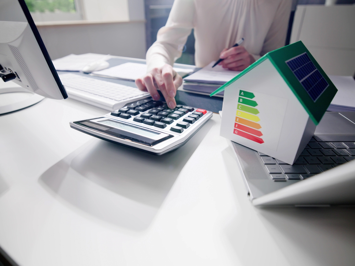 Johns Creek Energy-Efficient Home Remodeling: The Best Green Upgrades for Long-Term Savings 2 A person uses a calculator at a desk with a model house displaying an energy efficiency rating chart, next to a laptop—perfect for planning green upgrades or energy-efficient home remodeling in Johns Creek.