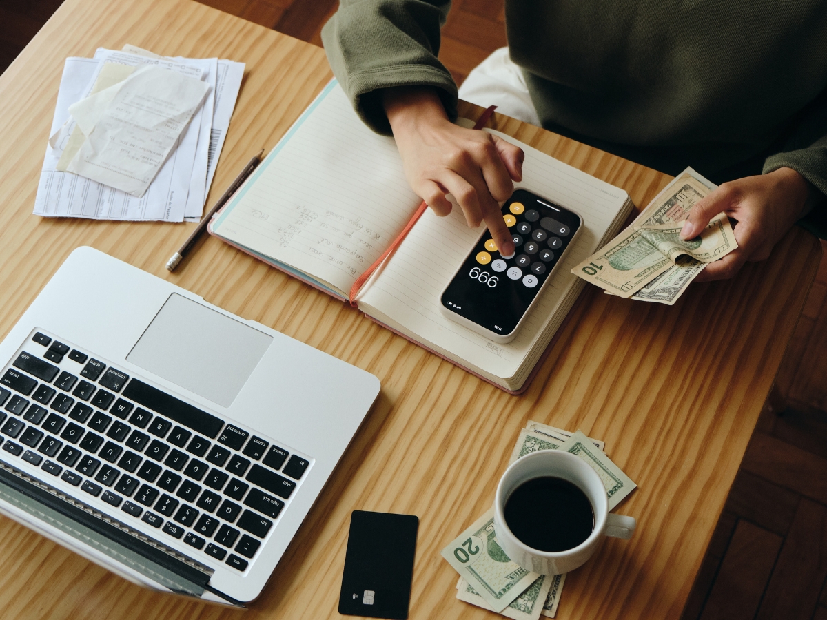 Johns Creek Energy-Efficient Home Remodeling: The Best Green Upgrades for Long-Term Savings 4 Person using a smartphone calculator, holding cash, with a notebook, laptop, receipts, and coffee cup on a wooden desk—planning energy-efficient home remodeling in Johns Creek with an eye on green upgrades.