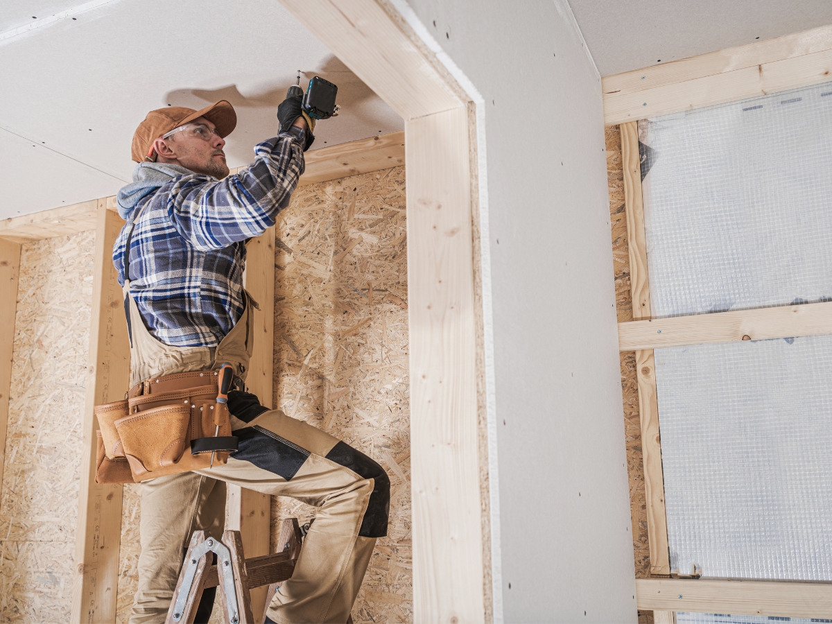 Johns Creek Energy-Efficient Home Remodeling: The Best Green Upgrades for Long-Term Savings 5 A construction worker on a ladder installs drywall on a ceiling inside a partially finished room with exposed wooden framing, part of an energy-efficient home remodeling project in Johns Creek.