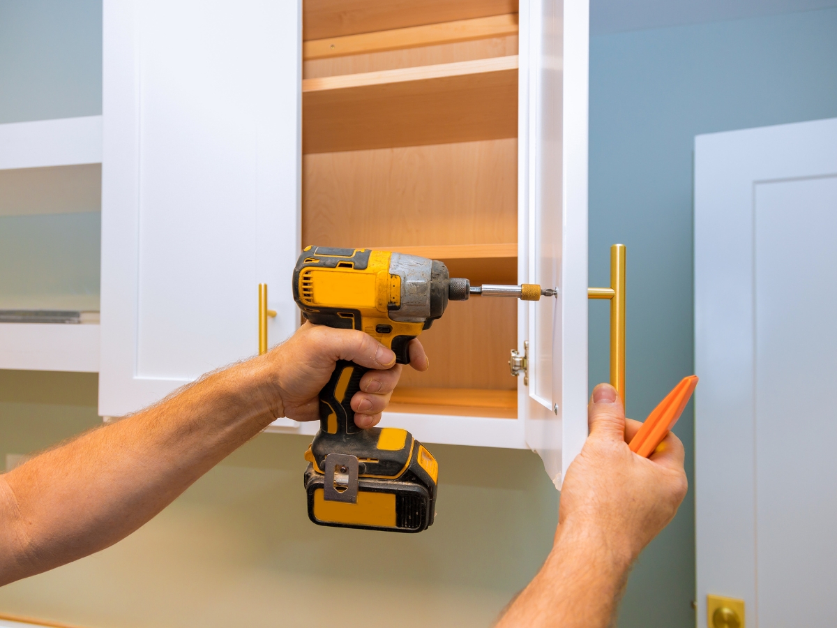 A person uses a cordless drill to install a gold handle on a white kitchen cabinet door, with the cabinet open and a plastic tool in the other hand—perfect details for Marietta Kitchen Remodeling projects.