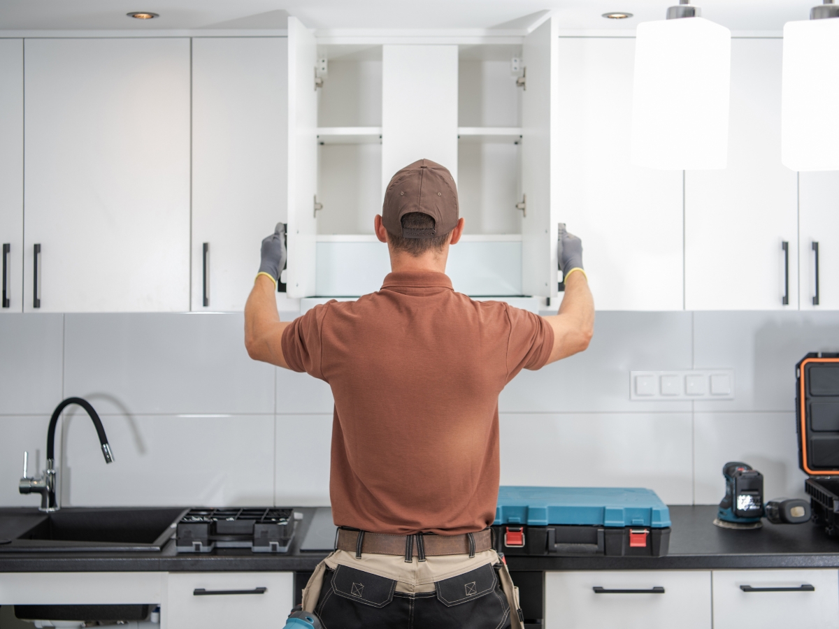 A worker in a brown shirt and cap installs or inspects an upper kitchen cabinet; tools and a toolbox are on the black countertop—another step in the Marietta Kitchen Remodeling process.