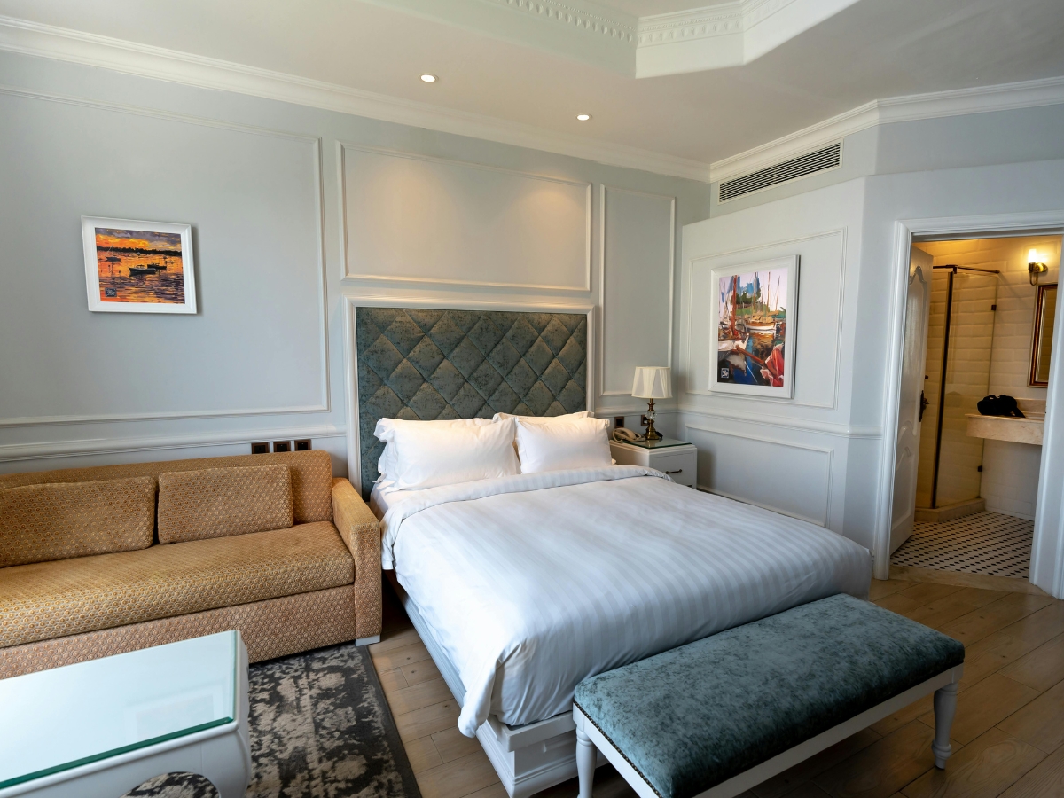 Roswell Basement Remodeling for Holiday Guests: Transform Your Space into a Comfortable Suite 2 A hotel room with a double bed, beige sofa, glass coffee table, padded bench, and two framed pictures on the wall—perfect inspiration for your Roswell Basement Remodeling for holiday guests. The bathroom is visible through an open door.