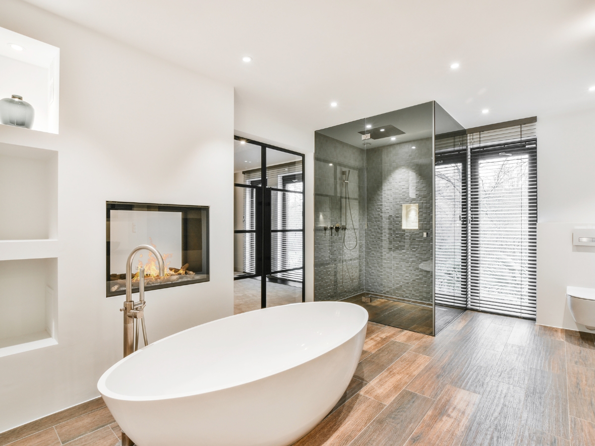 Modern bathroom with a freestanding bathtub, glass-enclosed shower, wall-mounted toilet, and wood-style flooring. Discover Sandy Plains GA Bathroom Remodeling Ideas featuring large windows with blinds and a built-in fireplace.