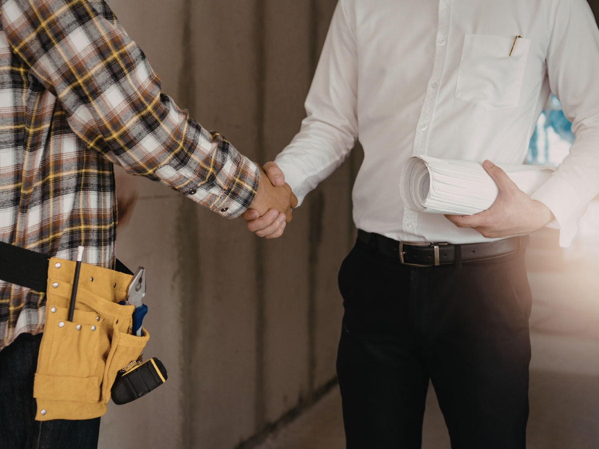 A General Contractor Checklist for Hiring the Right Professional For the Job 2 A person in a plaid shirt and tool belt shakes hands with another person in a white shirt holding rolled-up papers, likely reviewing a contractor checklist at a construction site.