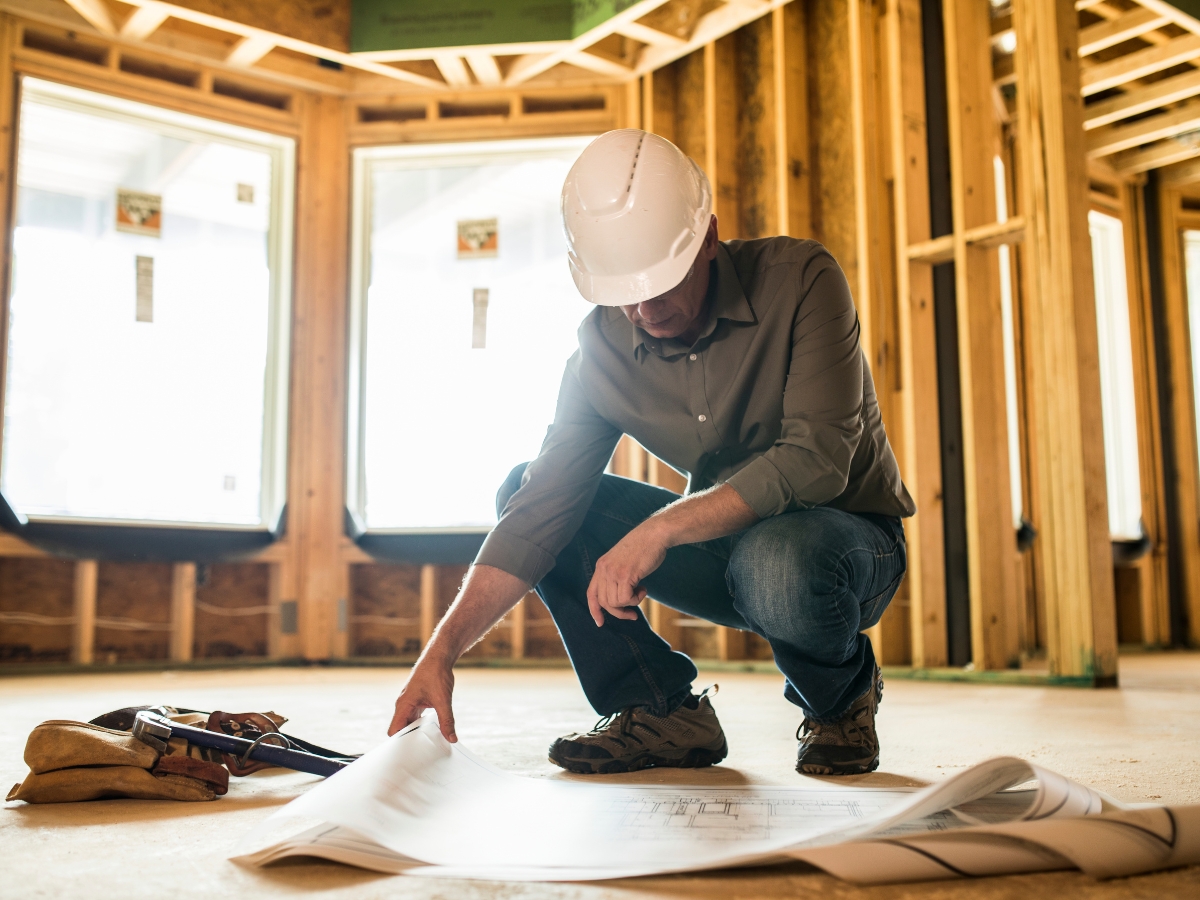 A General Contractor Checklist for Hiring the Right Professional For the Job 4 A person wearing a hard hat examines blueprints and a contractor checklist inside a building under construction with exposed wooden framing.