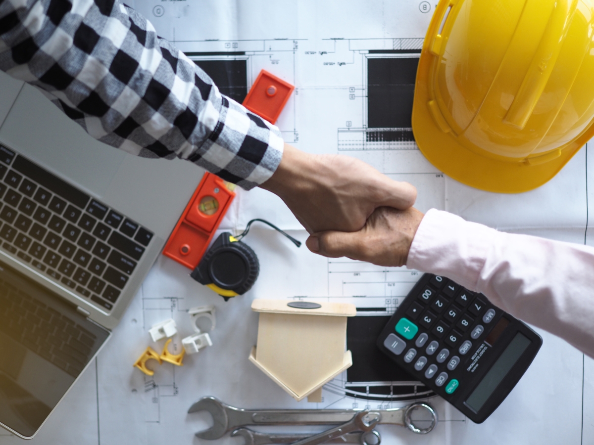 A General Contractor Checklist for Hiring the Right Professional For the Job 8 Two people shake hands over a desk with a laptop, calculator, construction tools, and a yellow hard hat on top of architectural blueprints, reviewing their contractor checklist.