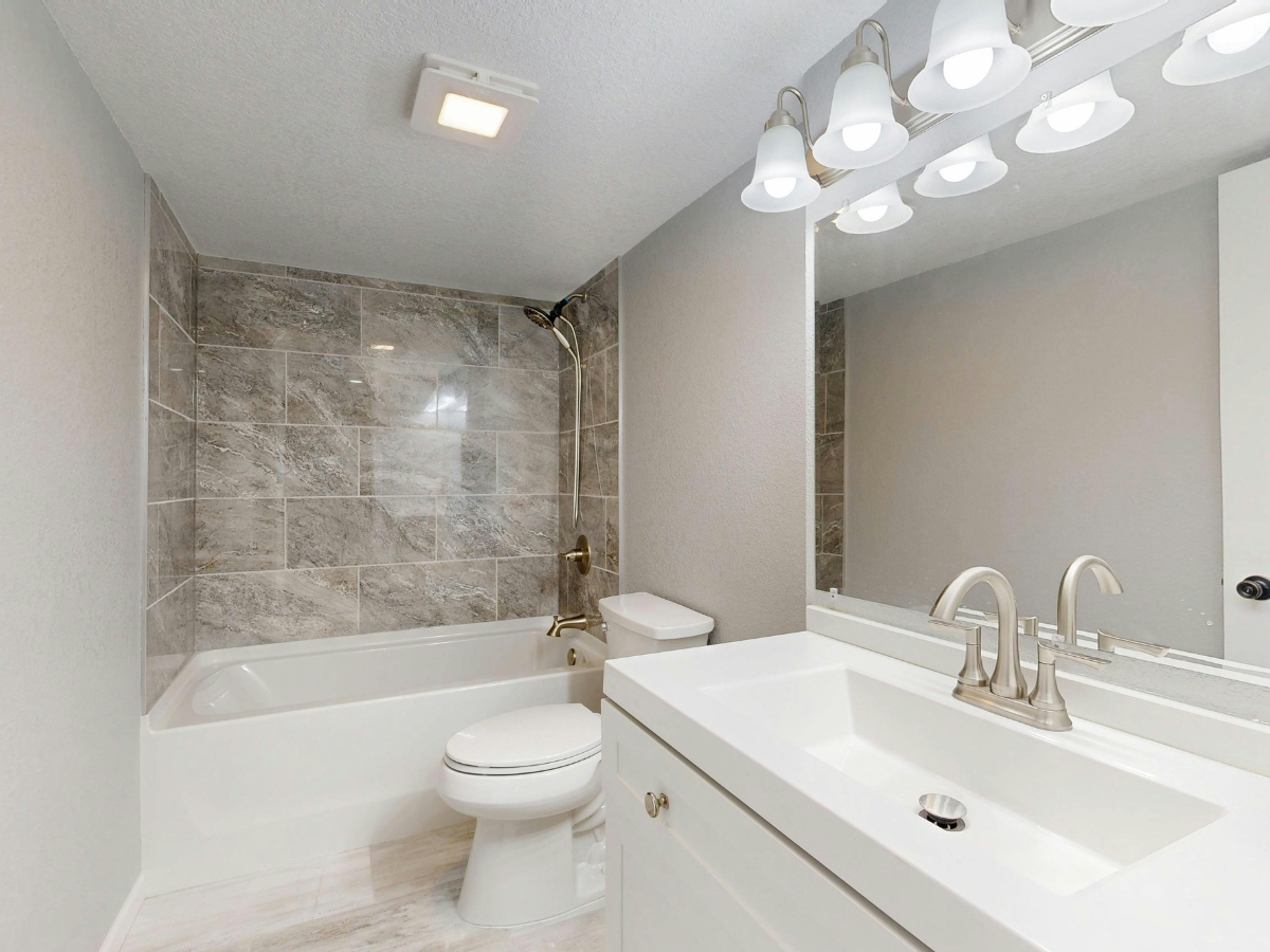 Modern bathroom with a white vanity, large mirror, toilet, and a bathtub-shower combo featuring gray stone tile surround. Ample lighting and bathroom ventilation keep the space bright and fresh.