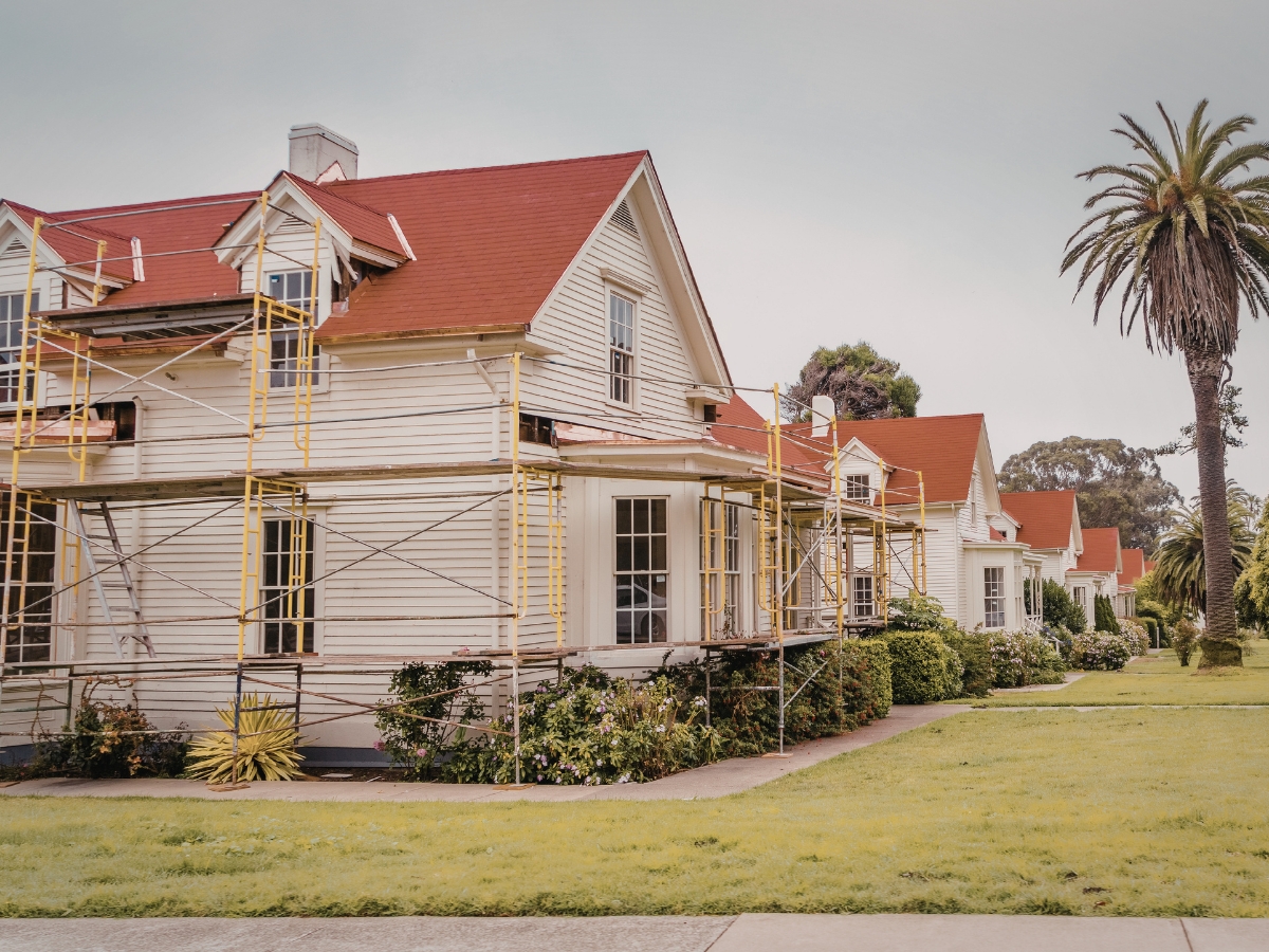 Two white historic homes with red roofs are surrounded by scaffolding, indicating renovation work. A palm tree and neatly cut grass are visible in the foreground.