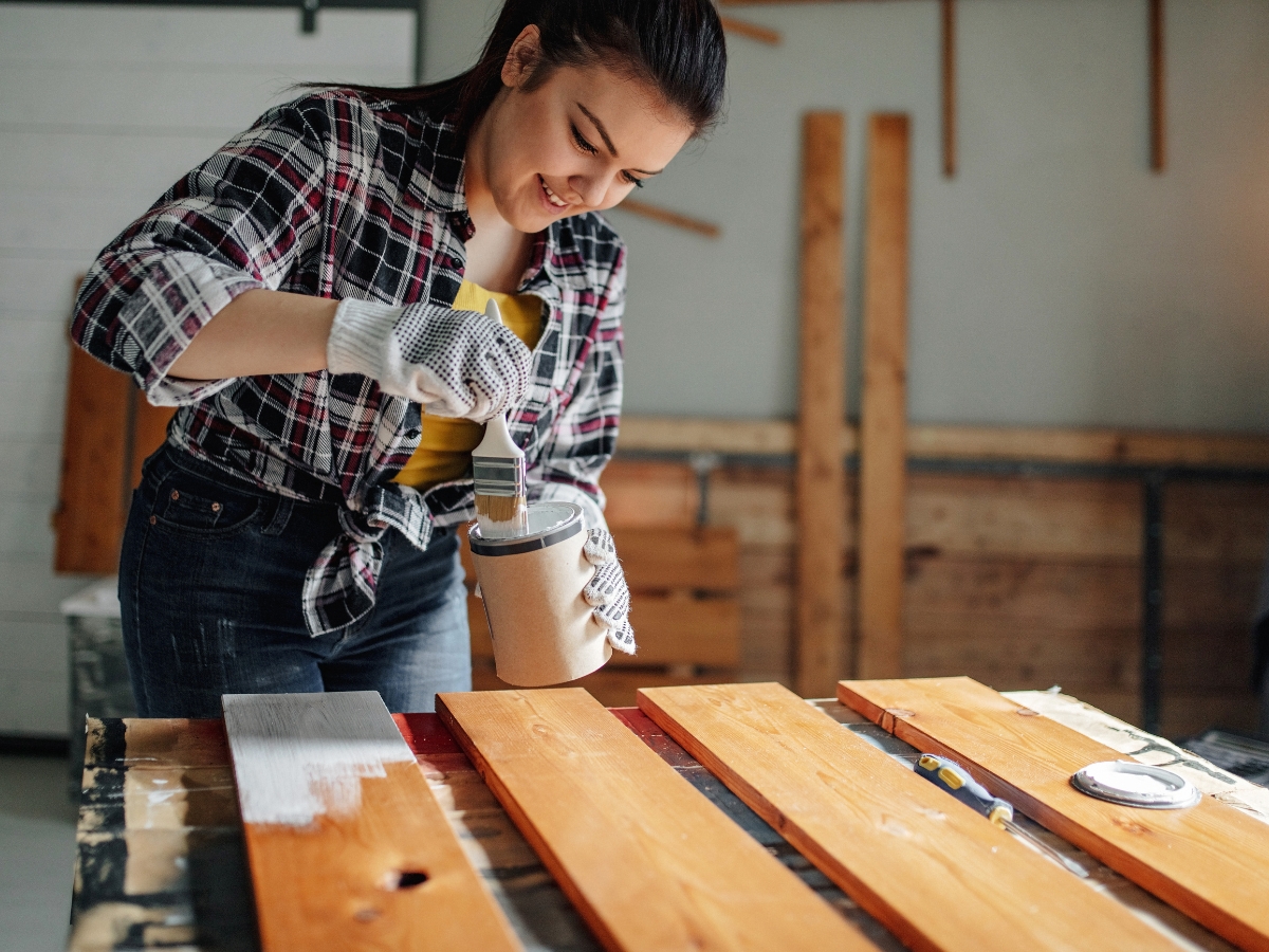 A person wearing gloves and a plaid shirt paints wooden boards white with a brush in a workshop, preparing materials for restoring historic homes.