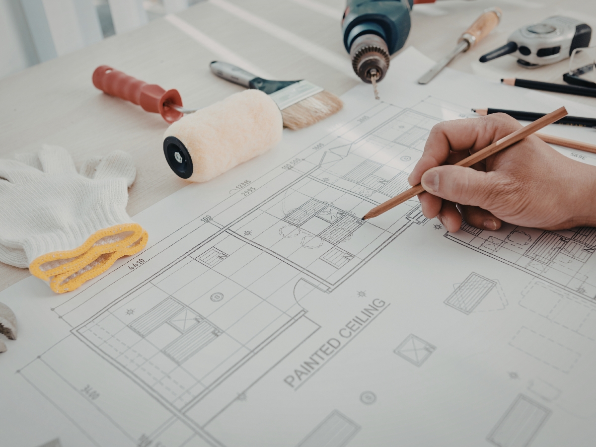 A hand draws on an architectural floor plan for historic homes, surrounded by tools including a paint roller, gloves, a drill, tape measure, and paintbrush.