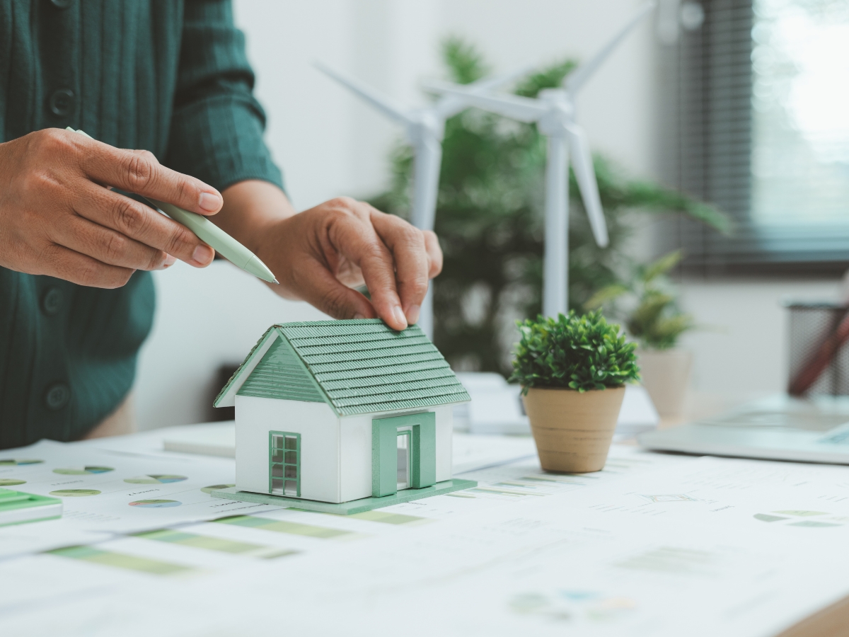 A person points a pen at a small model house—possibly inspired by historic homes—on a desk with papers, a potted plant, and wind turbine models in the background.