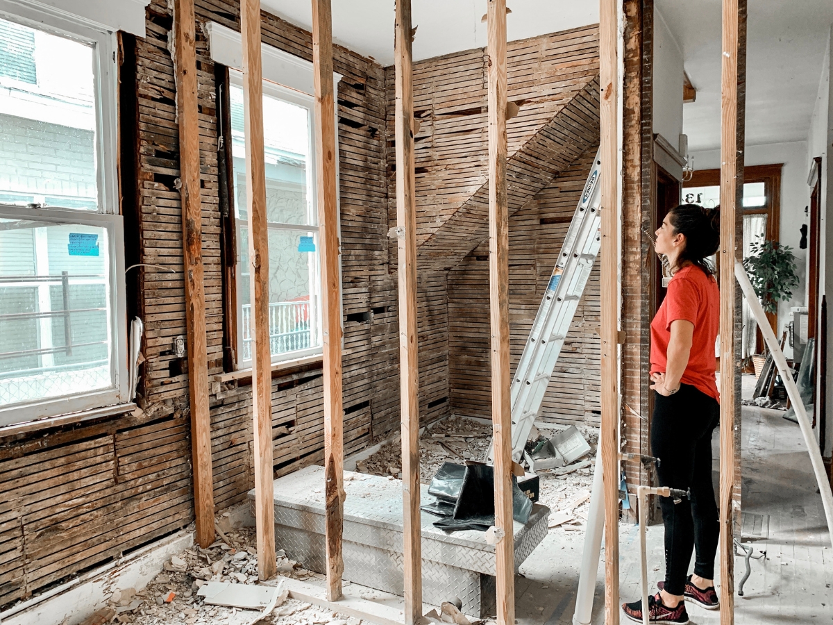A woman stands in a partially demolished room with exposed wall studs and debris on the floor, beginning the restoration of one of the area's historic homes; a ladder stands in the background, ready for use.