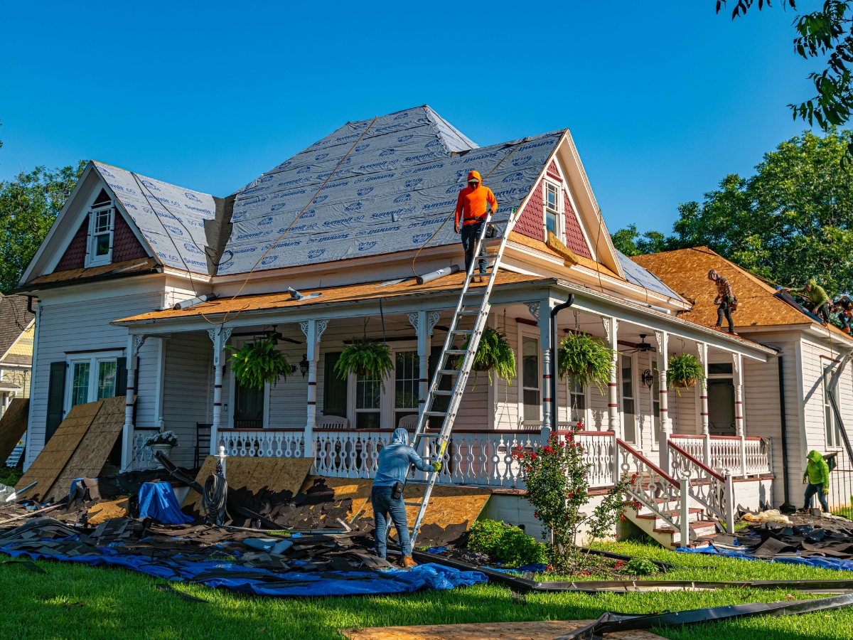 Workers replace the shingles on a large historic home's roof, with ladders, tools, and materials spread around the yard and porch on a sunny day.