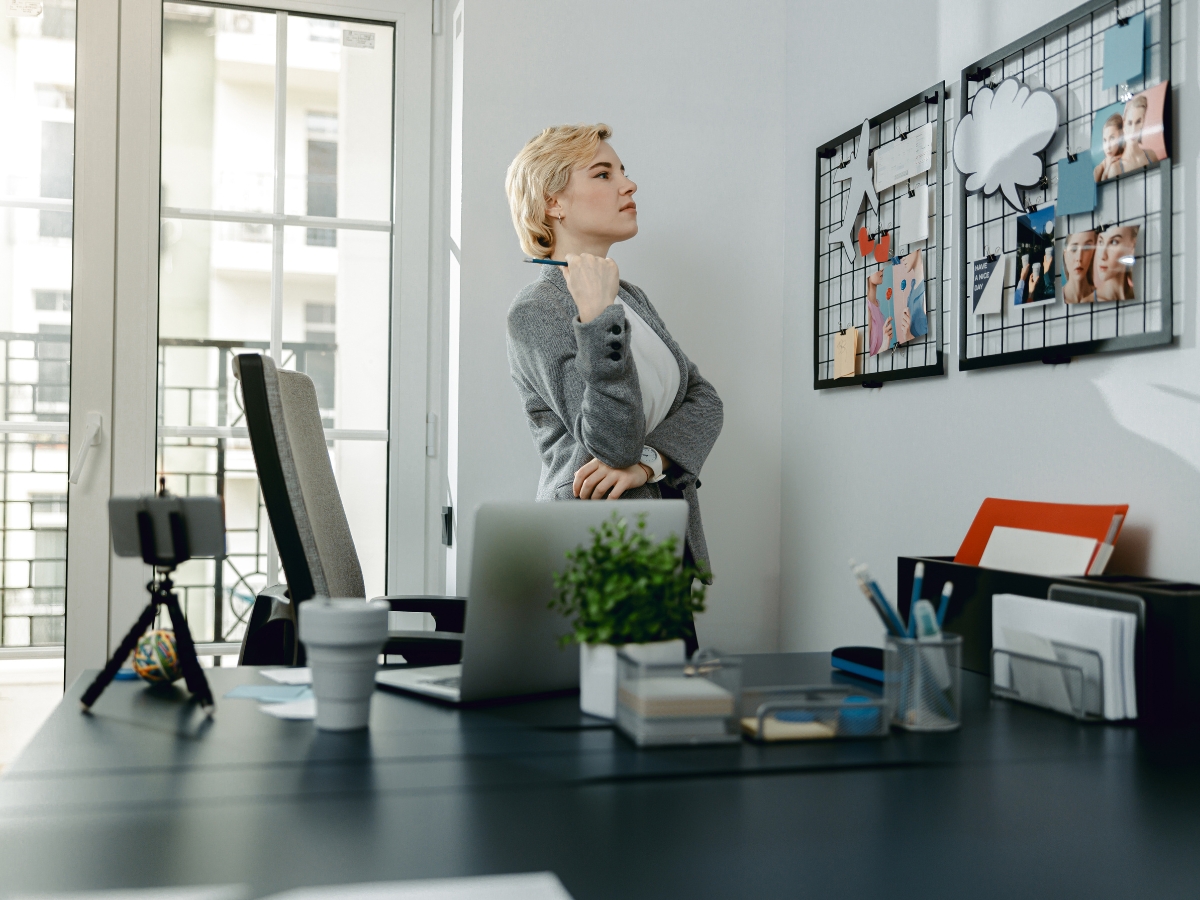 Person standing in an office, looking thoughtfully at a mood board filled with papers and images; a desk with a laptop, plant, and office supplies is in the foreground.