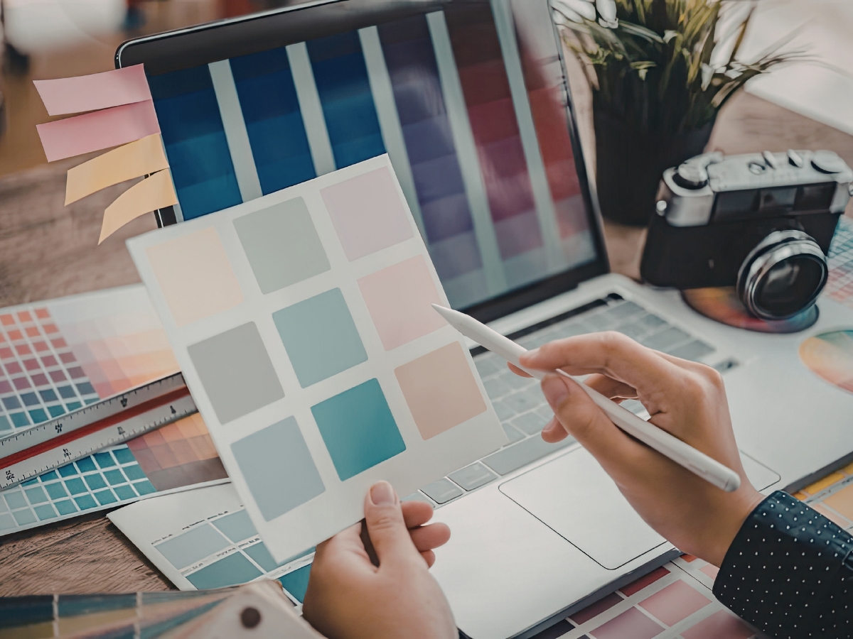A person compares color swatches in front of a laptop displaying a similar palette, with design tools, color charts, a mood board, and a camera on the desk.