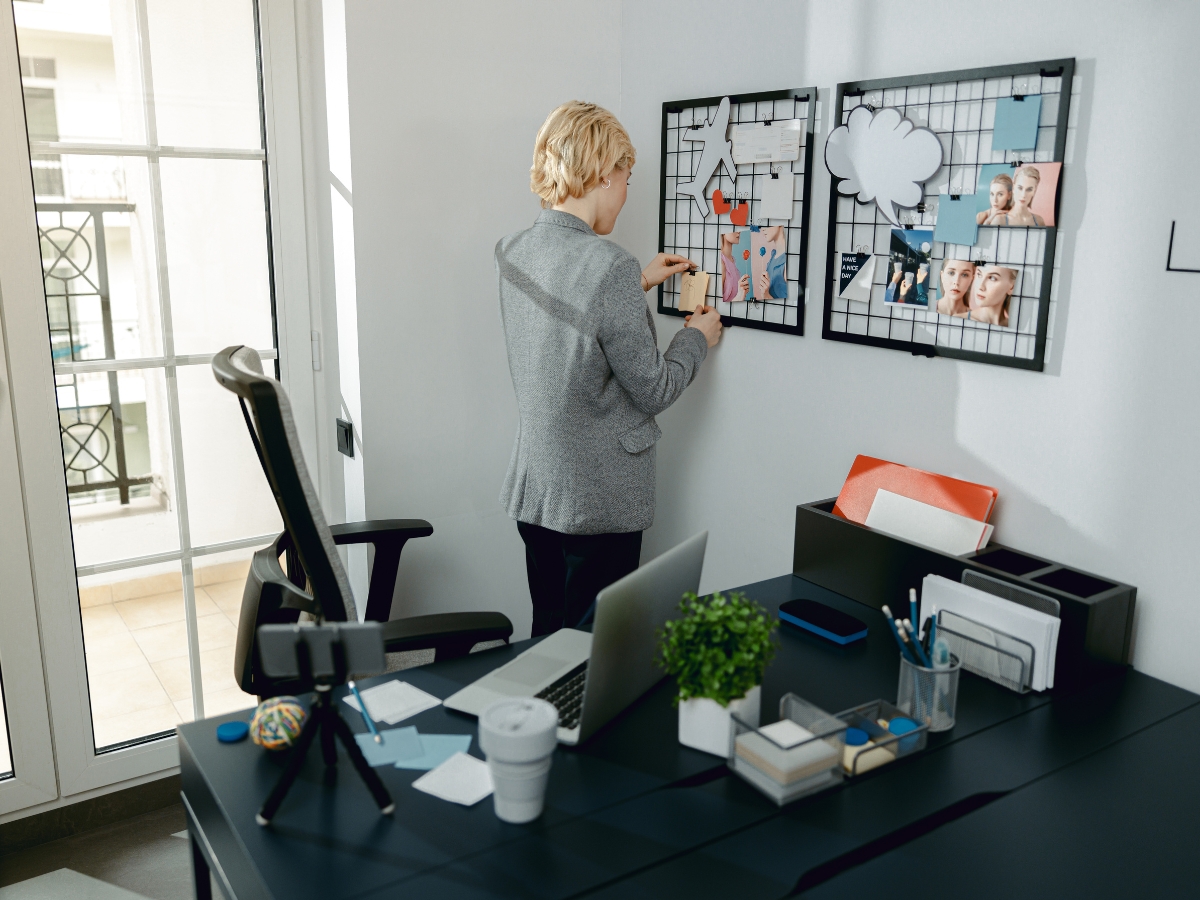 A person stands in an office pinning items to a wire grid mood board on the wall, with a desk in the foreground holding a laptop, coffee cup, and office supplies.