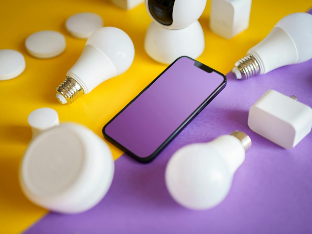 A smartphone with a purple screen is surrounded by smart lighting bulbs and plugs on a yellow and purple background.