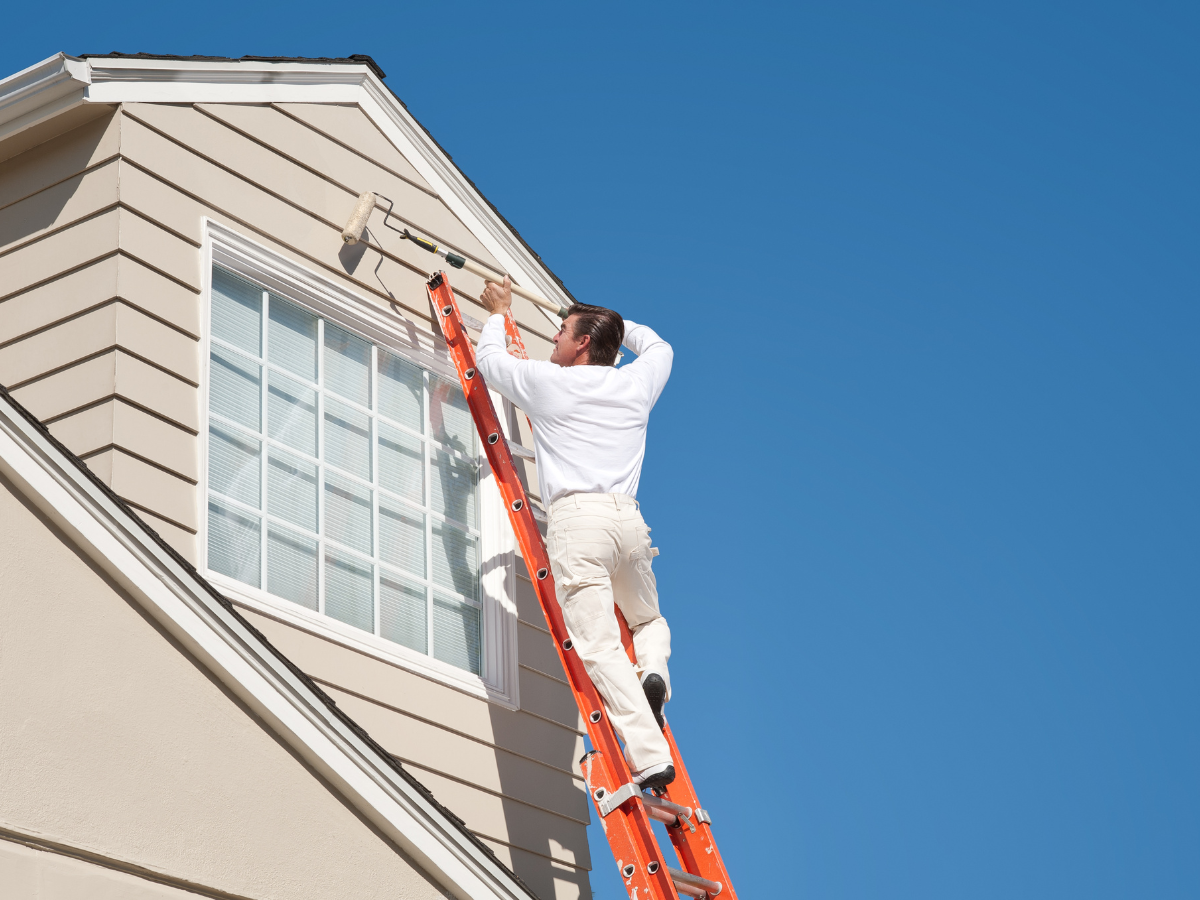 A person stands on an orange ladder applying exterior paint to the siding of a house with a roller, beneath a clear blue sky.
