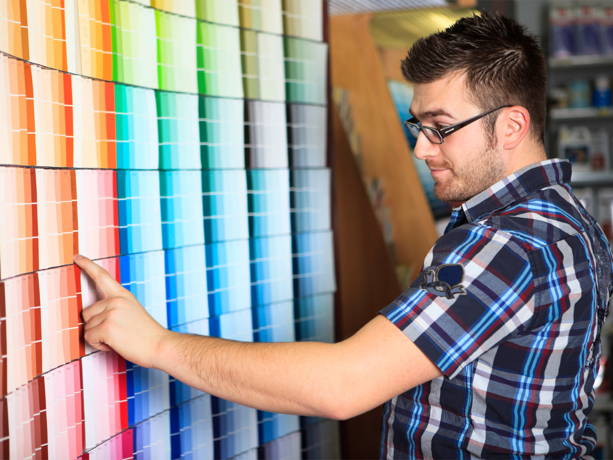 A man wearing glasses and a plaid shirt examines and points at exterior paint color swatches displayed on a wall in a store.