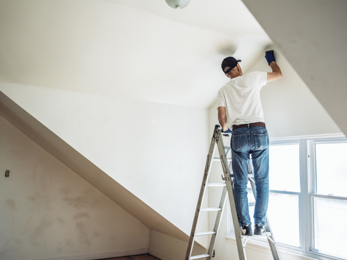 A person standing on a ladder applies exterior paint to the upper corner of a white room near a window, wearing jeans, a white t-shirt, and a cap.