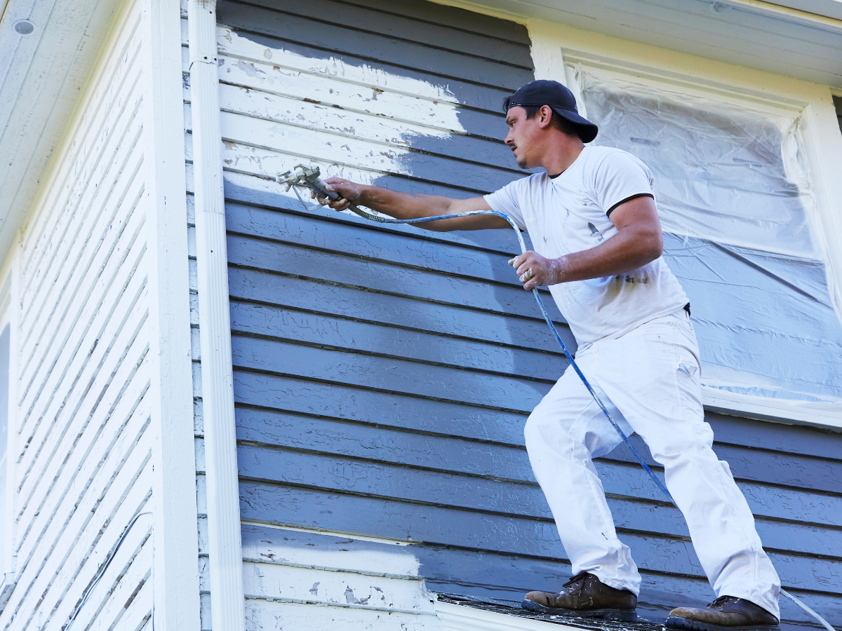 A man in white work clothes and a backwards cap uses a paint sprayer to apply fresh exterior paint, turning the siding of a house blue.