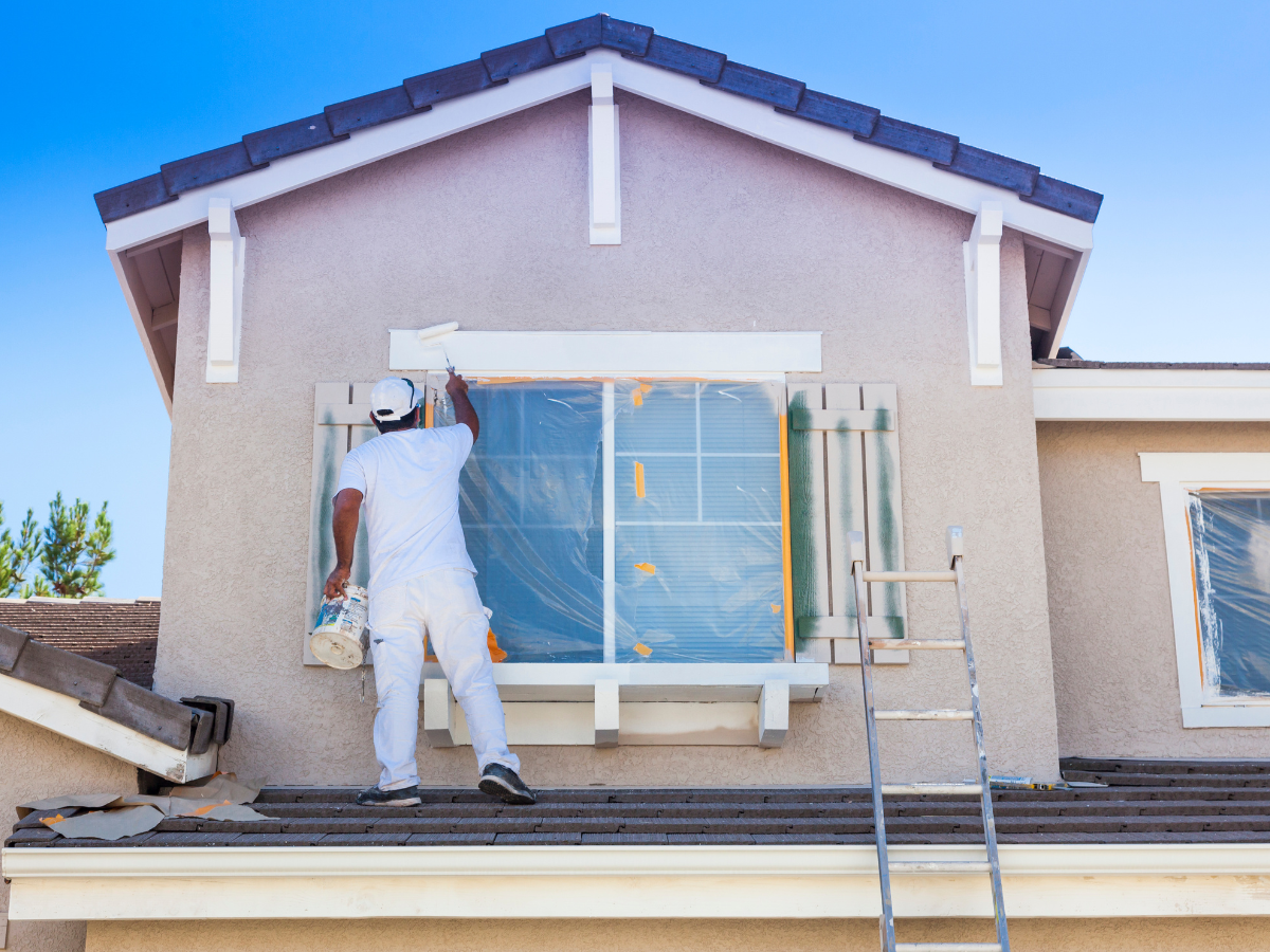 A person in white work clothes applies exterior paint to the trim around a second-story window, with a ladder nearby and plastic covering the window.