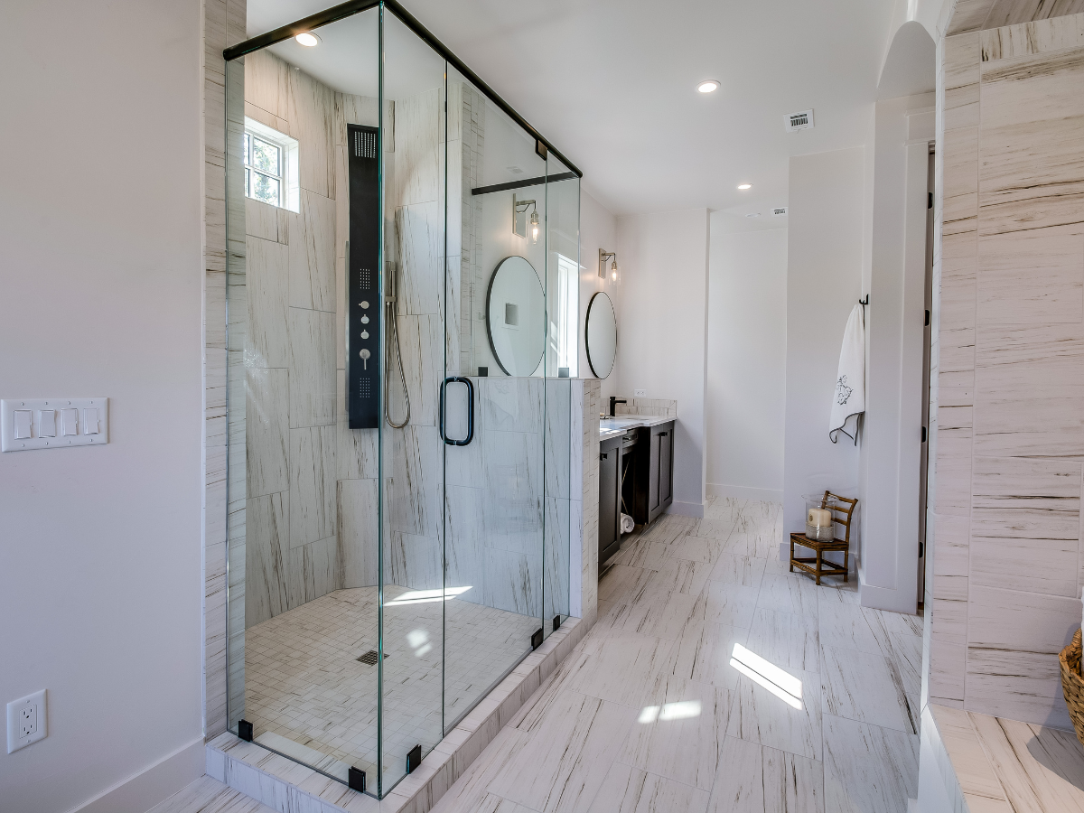Modern bathroom featuring a spacious walk-in shower with glass enclosure, double sink vanity with round mirrors, white tiled floor, and light walls.