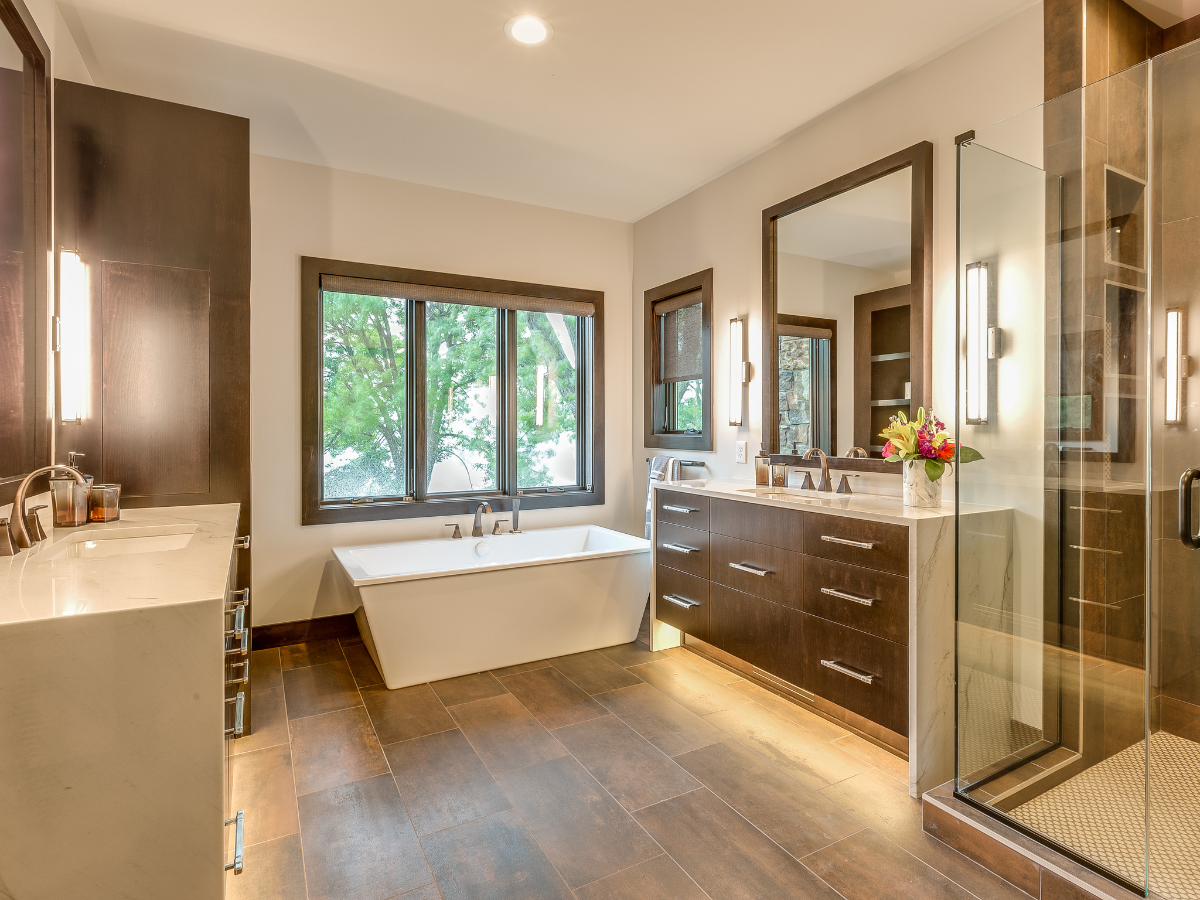 Modern bathroom with large windows, a freestanding bathtub, walk-in shower with glass doors, double sink vanity, dark wood cabinets, and brown tile flooring.