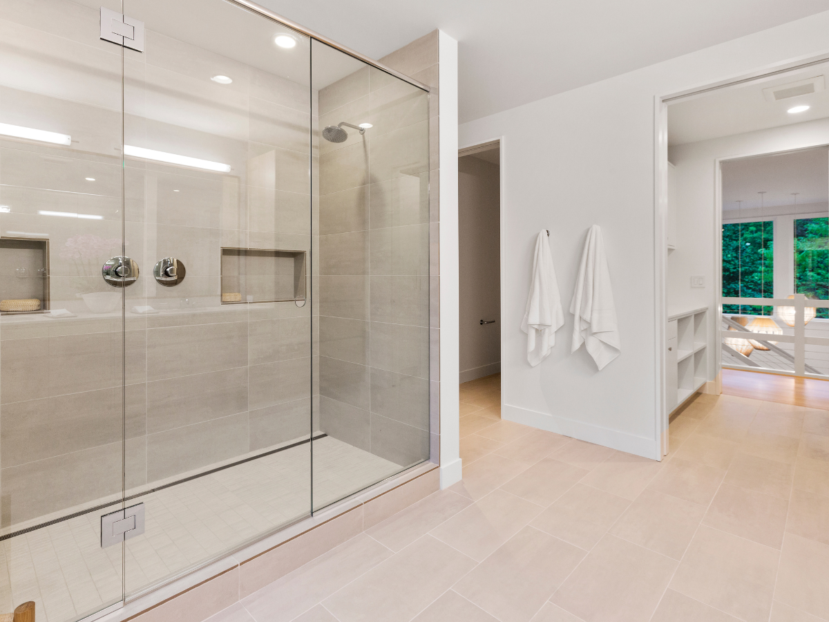 Spacious modern bathroom featuring a walk-in shower with glass enclosure, beige tile flooring, white walls, and two white towels hanging on the wall near a doorway.