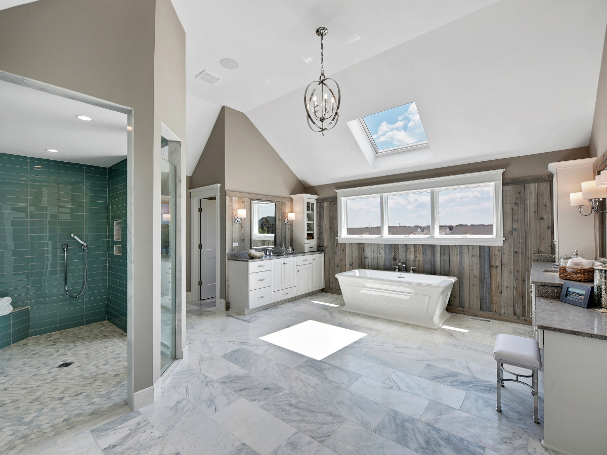 Spacious modern bathroom with a freestanding tub by large windows, double vanity, stylish walk-in shower with green tiles, marble floor, and a skylight.