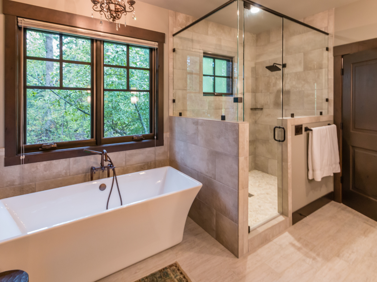 Modern bathroom with a freestanding white tub, a walk-in shower enclosed in glass, large windows offering a forest view, and beige tile flooring and walls.