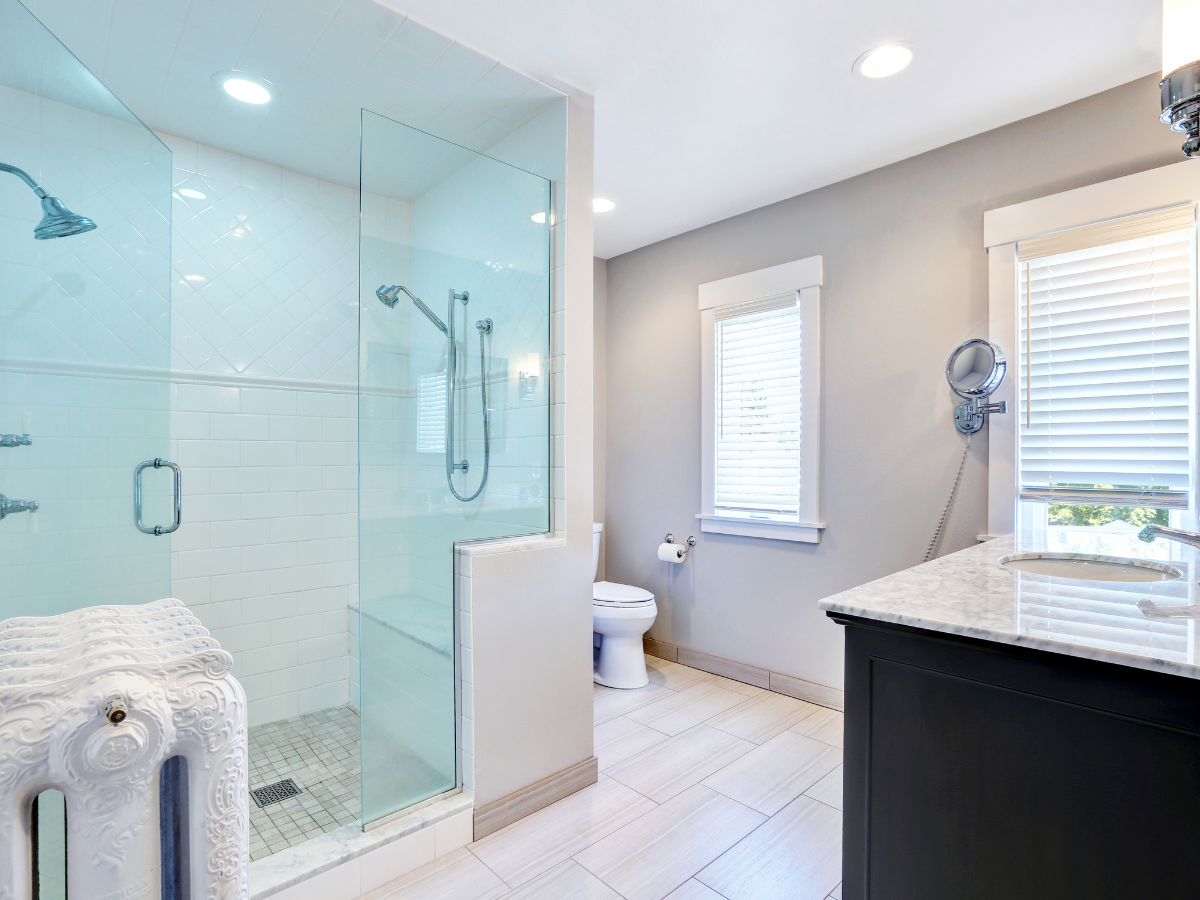Modern bathroom with walk-in shower featuring a glass enclosure, toilet, vanity with marble countertop, wall mirror, and two windows with blinds.