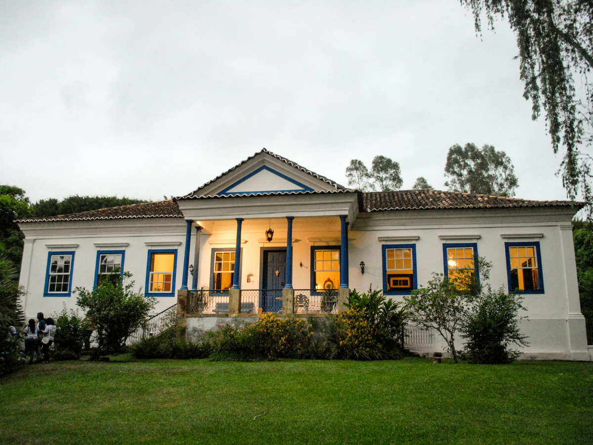 Effective Lighting Design for Home Remodeling 5 A white colonial-style house with blue trim features thoughtfully crafted lighting design, illuminating its windows and central front porch, all framed by a green lawn and shrubs on a cloudy day.
