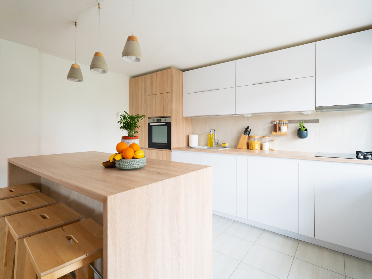 Achieving a Minimalist Design in Your Home Remodel 4 Modern kitchen with light wood cabinets, a central island with stools, built-in oven, and pendant lights. Showcasing minimalist design, the space features a bowl of oranges and a potted plant on the island.