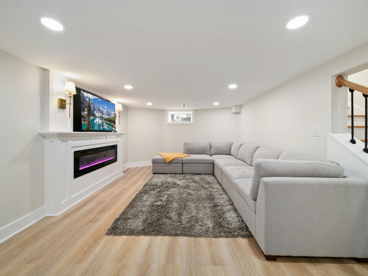 Achieving a Minimalist Design in Your Home Remodel 6 Modern basement living room with a minimalist design featuring a gray sectional sofa, wall-mounted TV above a fireplace, shag rug, light wood floors, and recessed lighting.