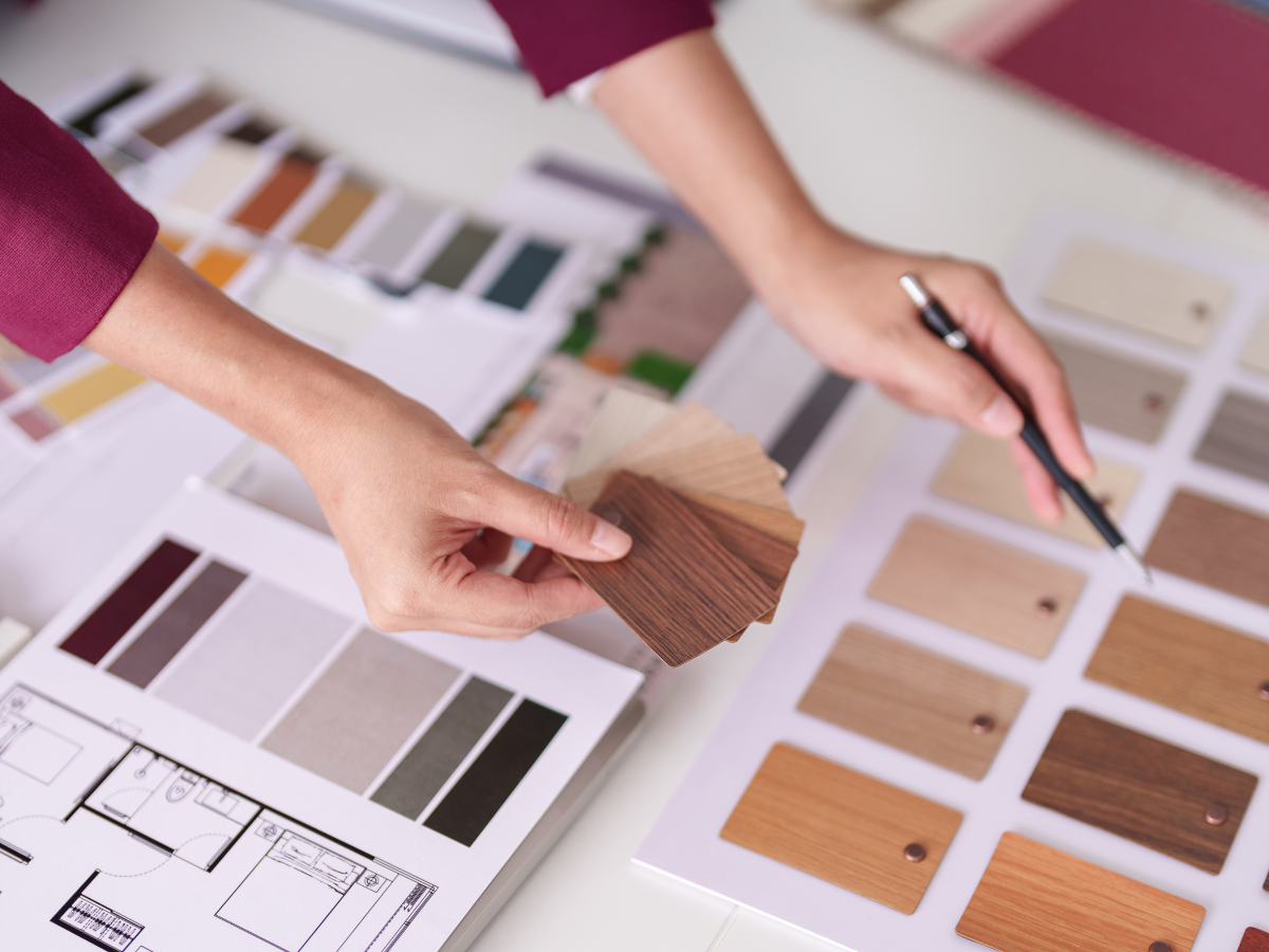 Achieving a Minimalist Design in Your Home Remodel 7 Person selecting wood veneer samples over architectural plans and color swatches, holding a pen and comparing finishes on a minimalist design workspace.