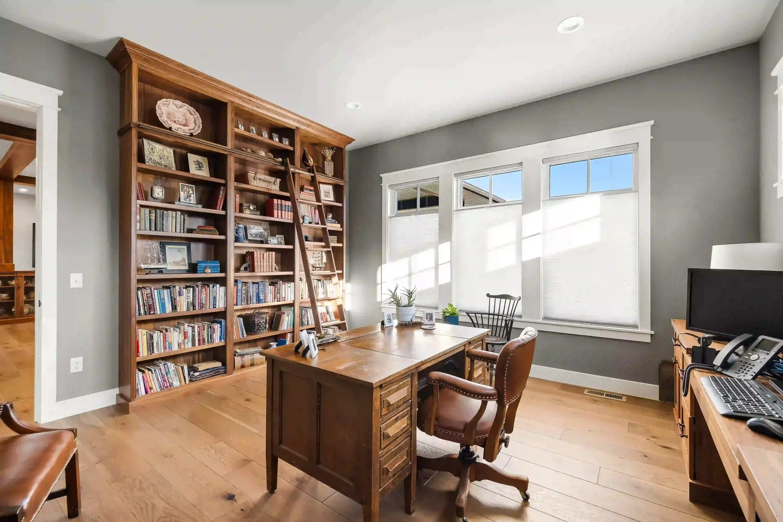 A home office with a wooden desk, leather chairs, a large bookshelf filled with books, hardwood floors, and windows letting in natural light.