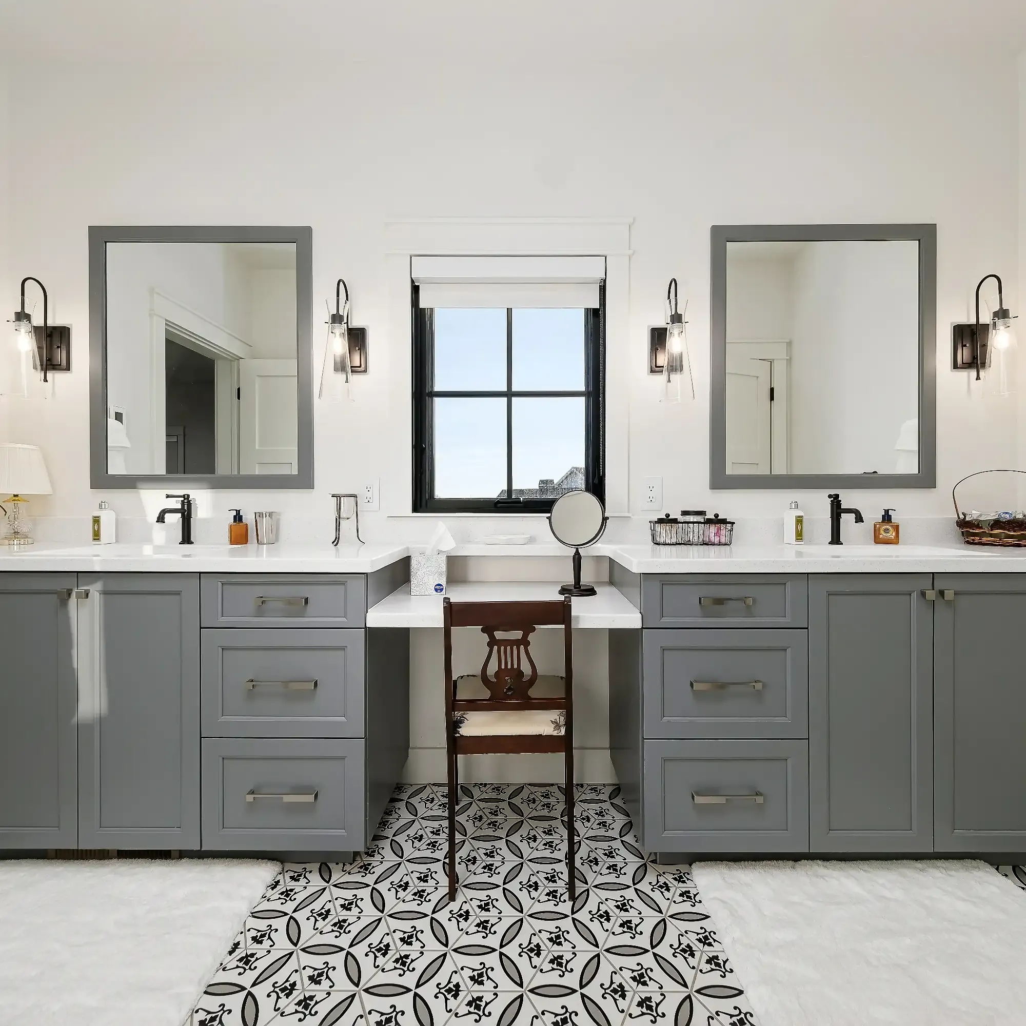 Modern bathroom with gray cabinets, double sinks, two mirrors, a makeup vanity in the center, black window, wall sconces, and patterned tile floor.