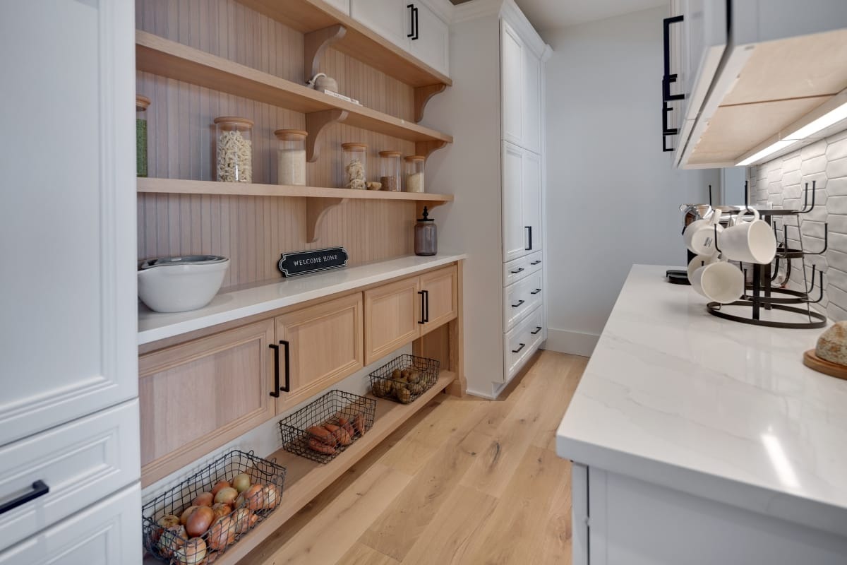 A pantry with wooden shelves holding jars of dry goods, baskets of produce below, white cabinets, and a counter with mugs hanging on a rack.