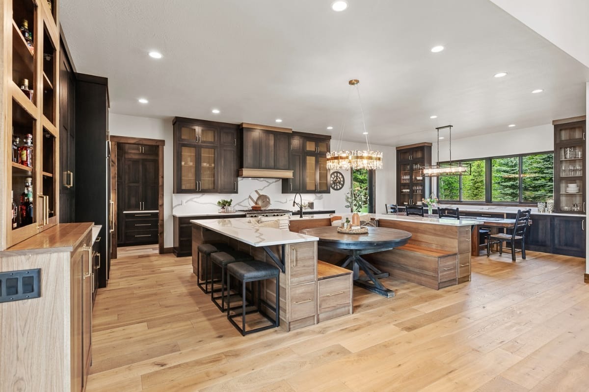 Modern kitchen with dark wood cabinets, large island with barstools, built-in bench seating around a round table, and wide plank light wood flooring. Large windows show green trees outside.
