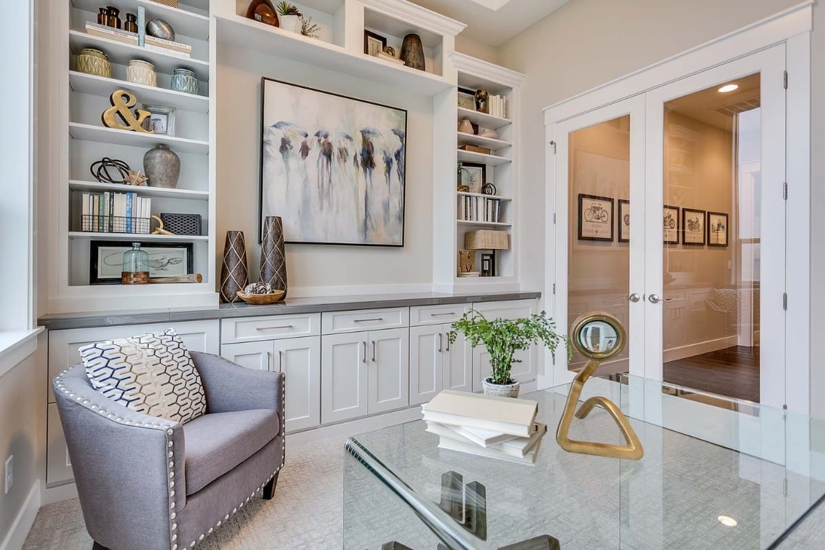 Modern home office with built-in shelves, abstract wall art, glass desk, decorative items, gray armchair, books, and double glass doors.