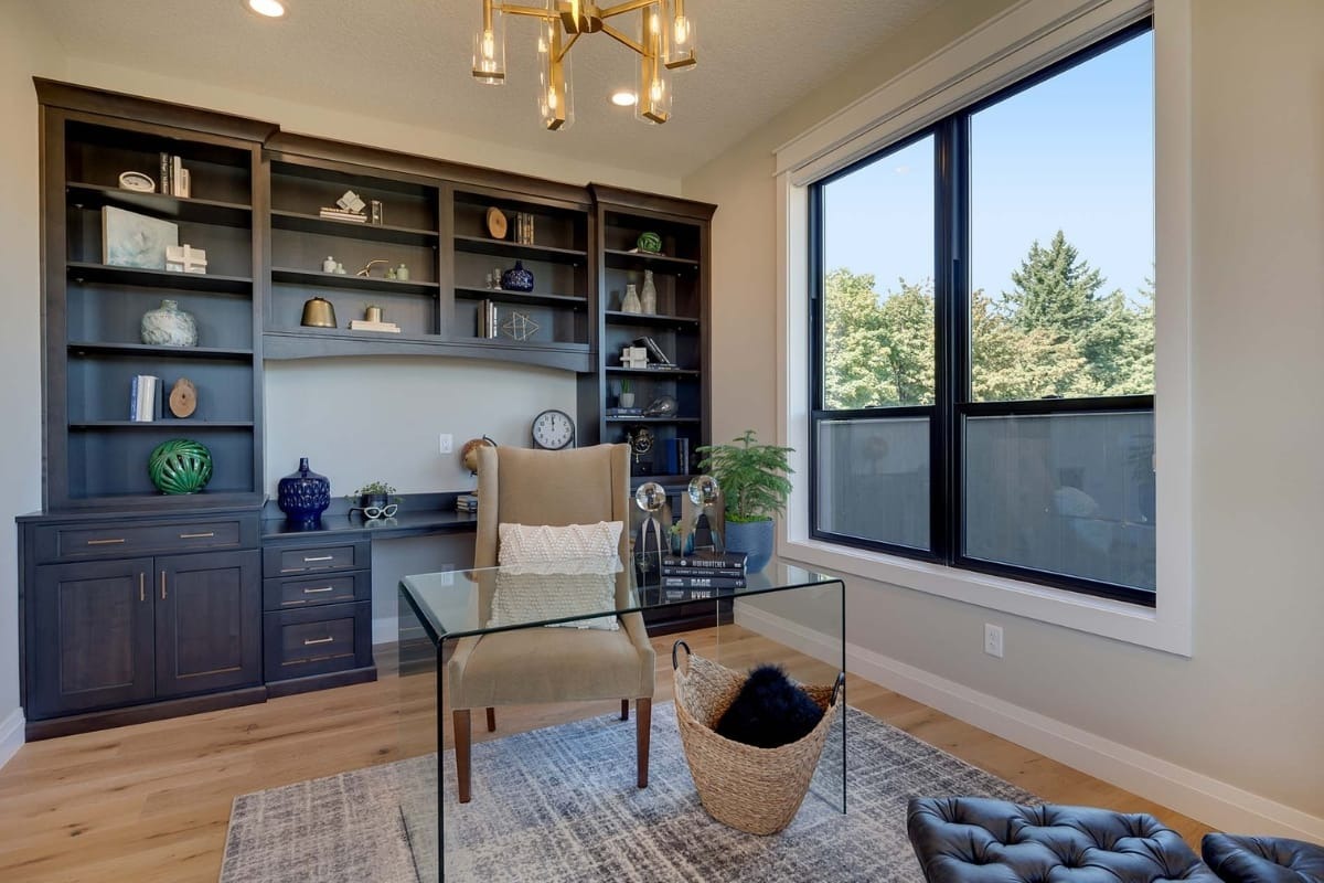 Modern home office with a glass desk, beige chair, built-in dark wood shelves, large window, and decorative items on the shelves and floor.