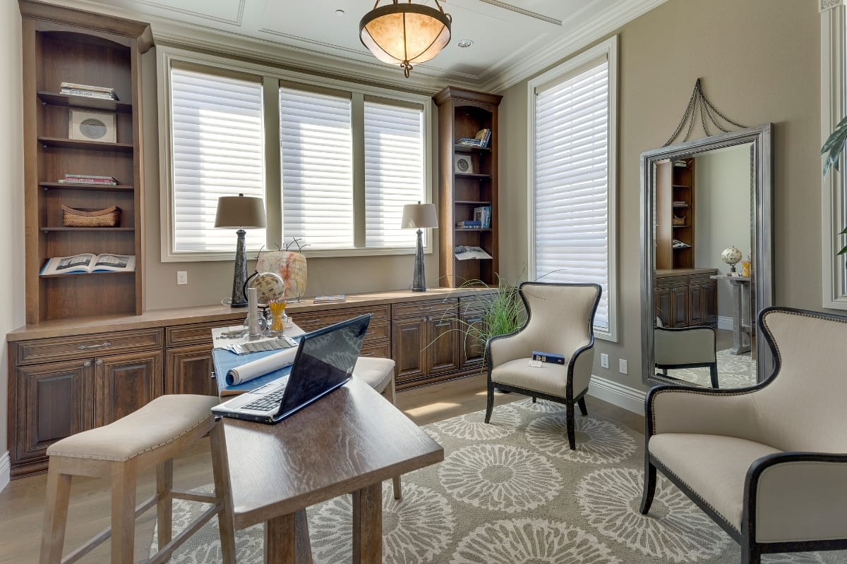 Home office with a wooden desk, open laptop, two armchairs, built-in shelves, large mirror, and windows with blinds. Neutral colors and patterned rug.