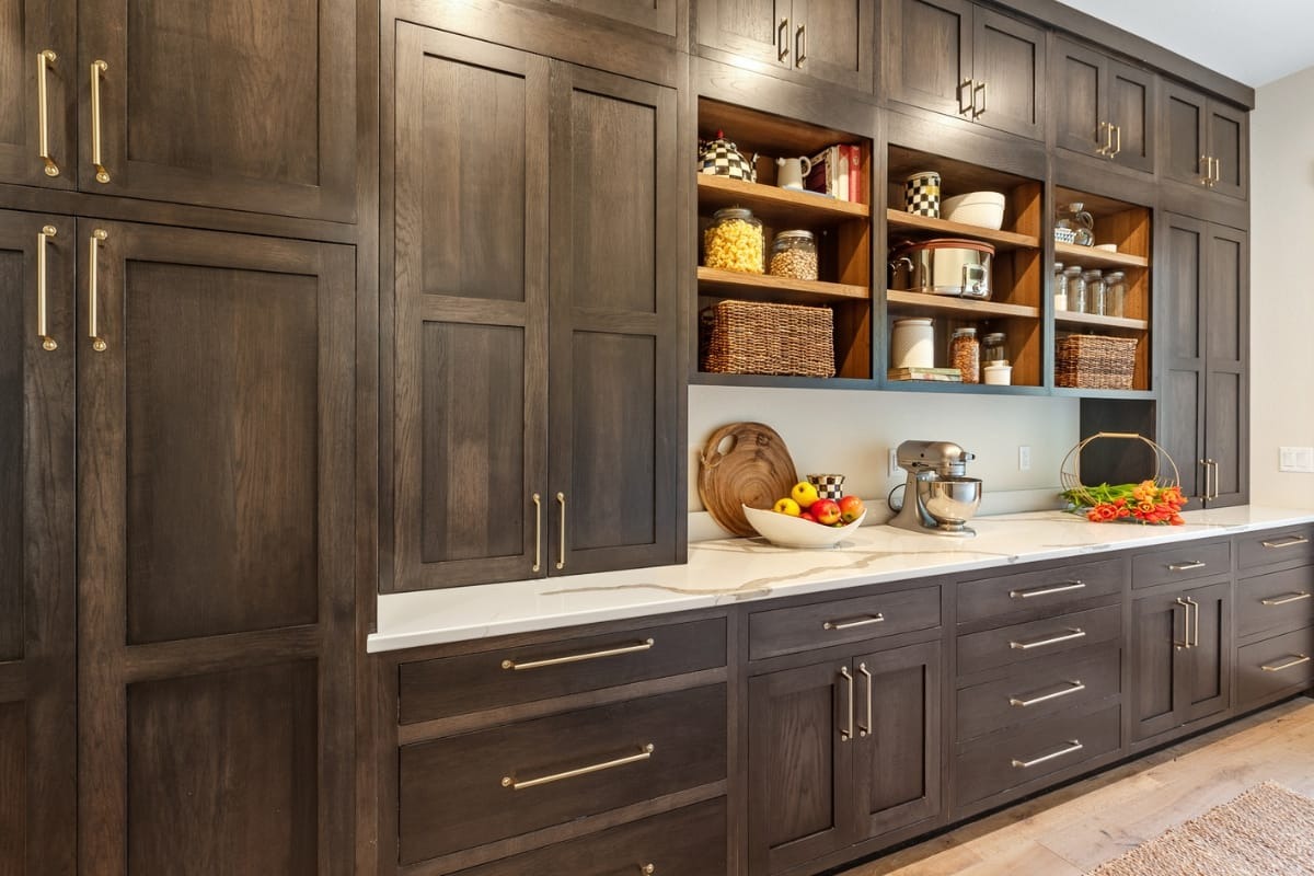 Modern kitchen with dark wood cabinets, white countertop, open shelves holding jars and baskets, and small appliances including a stand mixer and cutting boards.