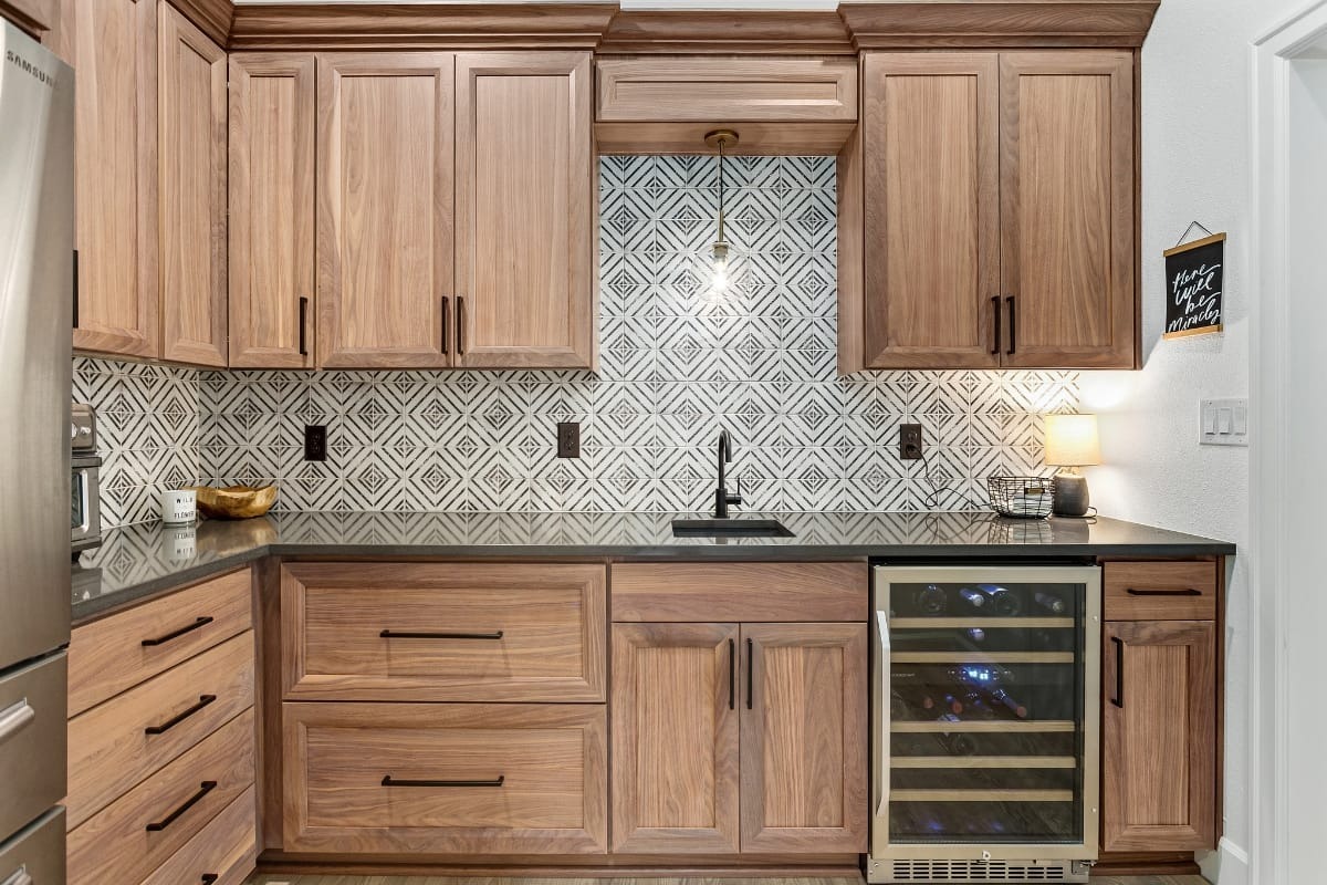 Modern kitchen with wood cabinets, geometric tile backsplash, black countertop, small sink, wine fridge, and under-cabinet lights. A lamp and small chalkboard are on the right countertop.
